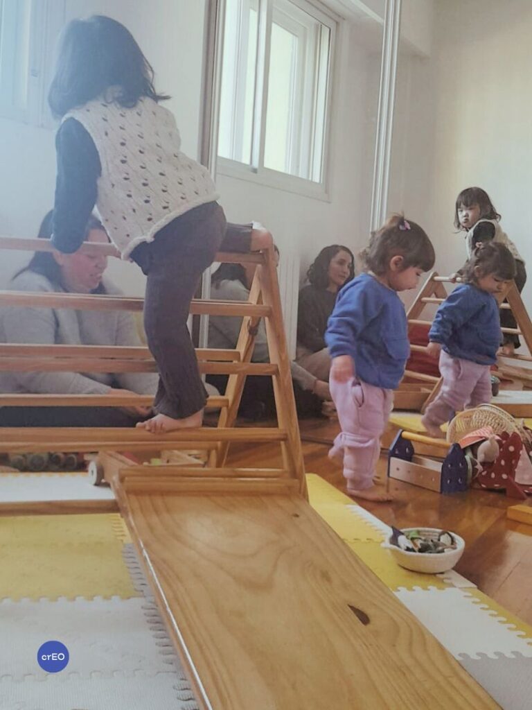 Niña pequeña trepando triángulo Pikler en espacio de juego libre mientras otros bebés juegan alrededor y adultos observan.