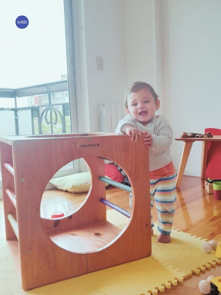 Bebé de pie agarrado del cubo Pikler que utiliza como sostén para mantenerse de pie en un espacio de movimiento y juego libre.